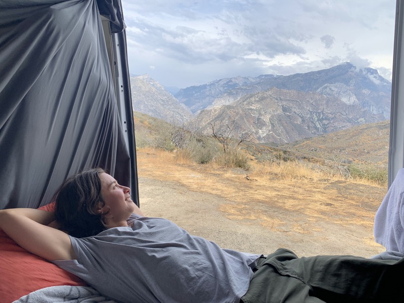 Rey lying down on a mattress in a van with the side door open, looking out on a mountain landscape