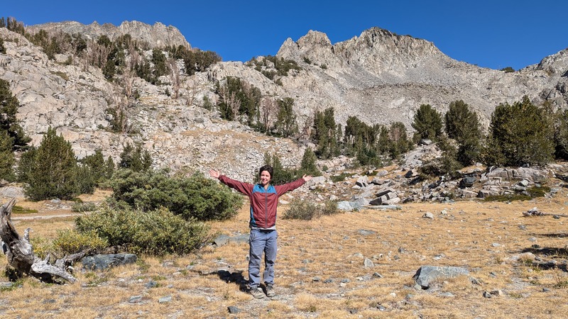 Rey in a grassy field in the mountains, arms raised and smiling