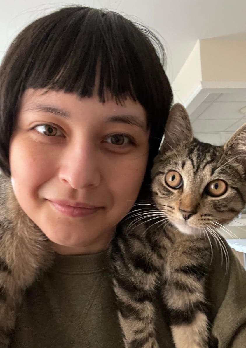 Lily with shortish straight black hair, smiling at the camera, with a very cute cat with stripes and orange eyes