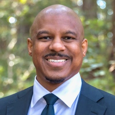 A photo of Chris Musei-Sequeria, a Black man, wearing a suit and tie and smiling at the camera.