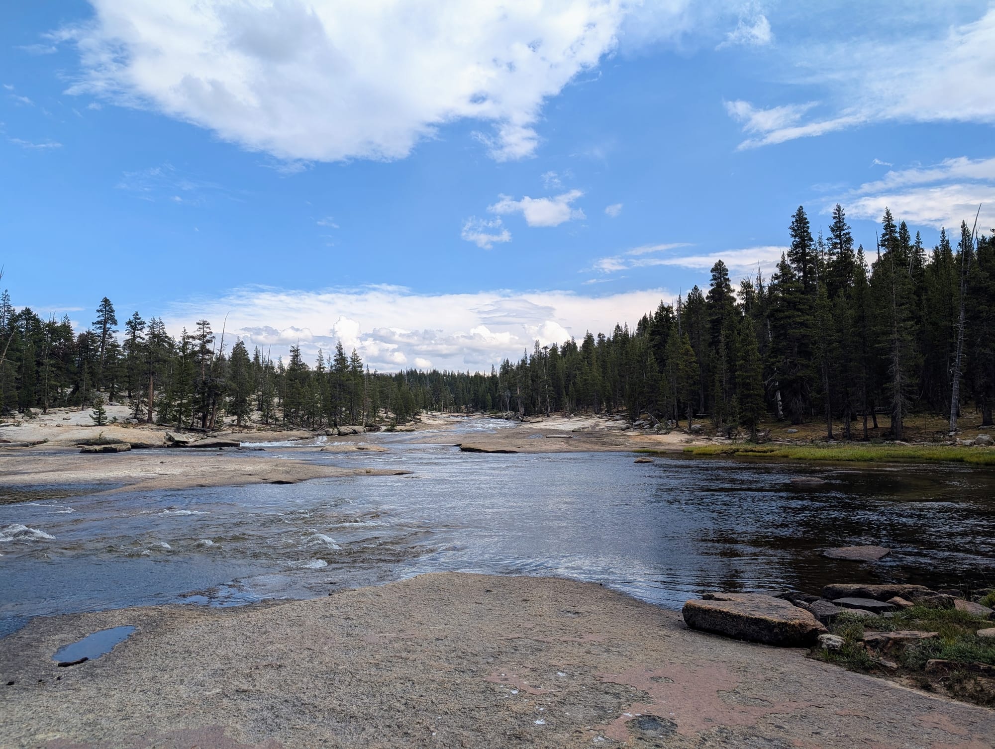 Water flowing over a large, flat granite area, with evergreen trees in the background and the sky reflected in the water