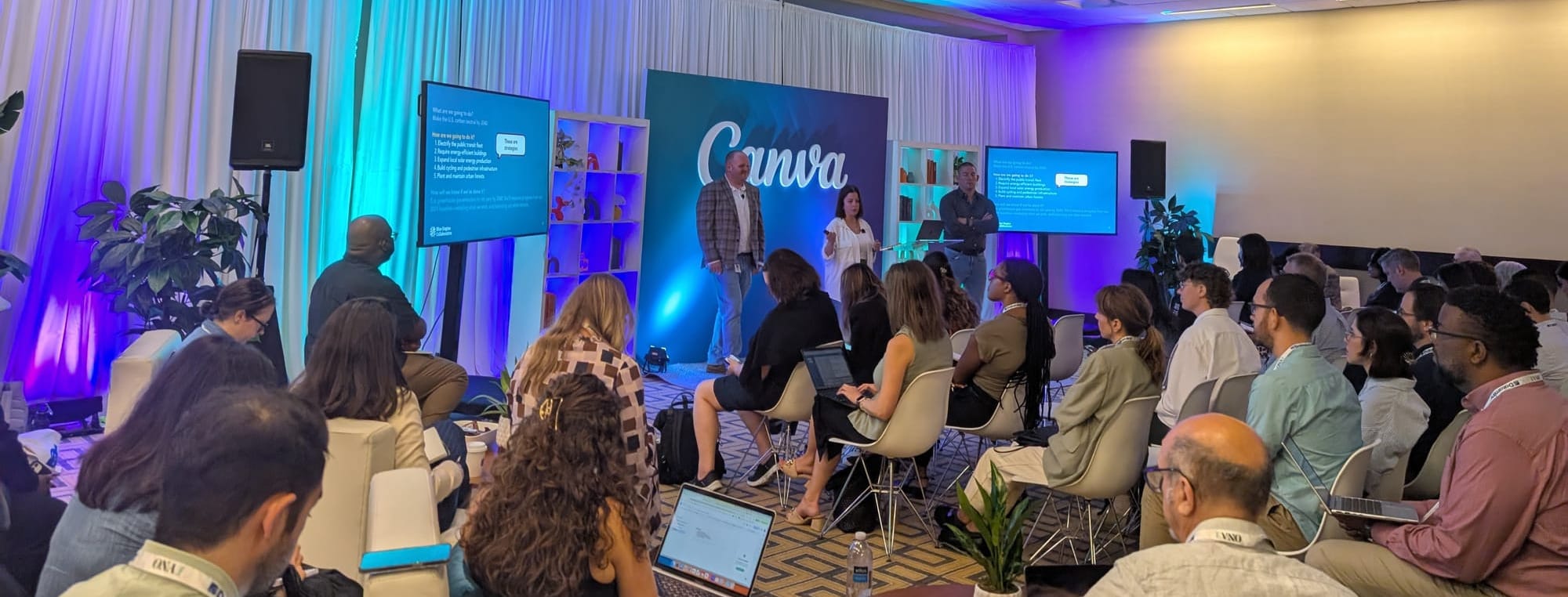 A room full of people learns from three people standing in front of a "Canva" backdrop and a slide presentation