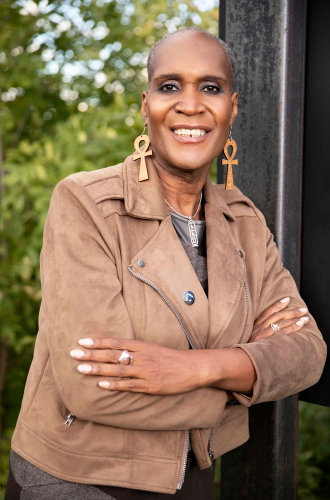 A photo of Andrea Jenkins, a Black woman with very short hair, eyeshadow, large earrings in the shape of a cross with a loop on top (this shape is the Ankh from Egypt), and a brown suede jacket, crossing her arms and smiling at the camera
