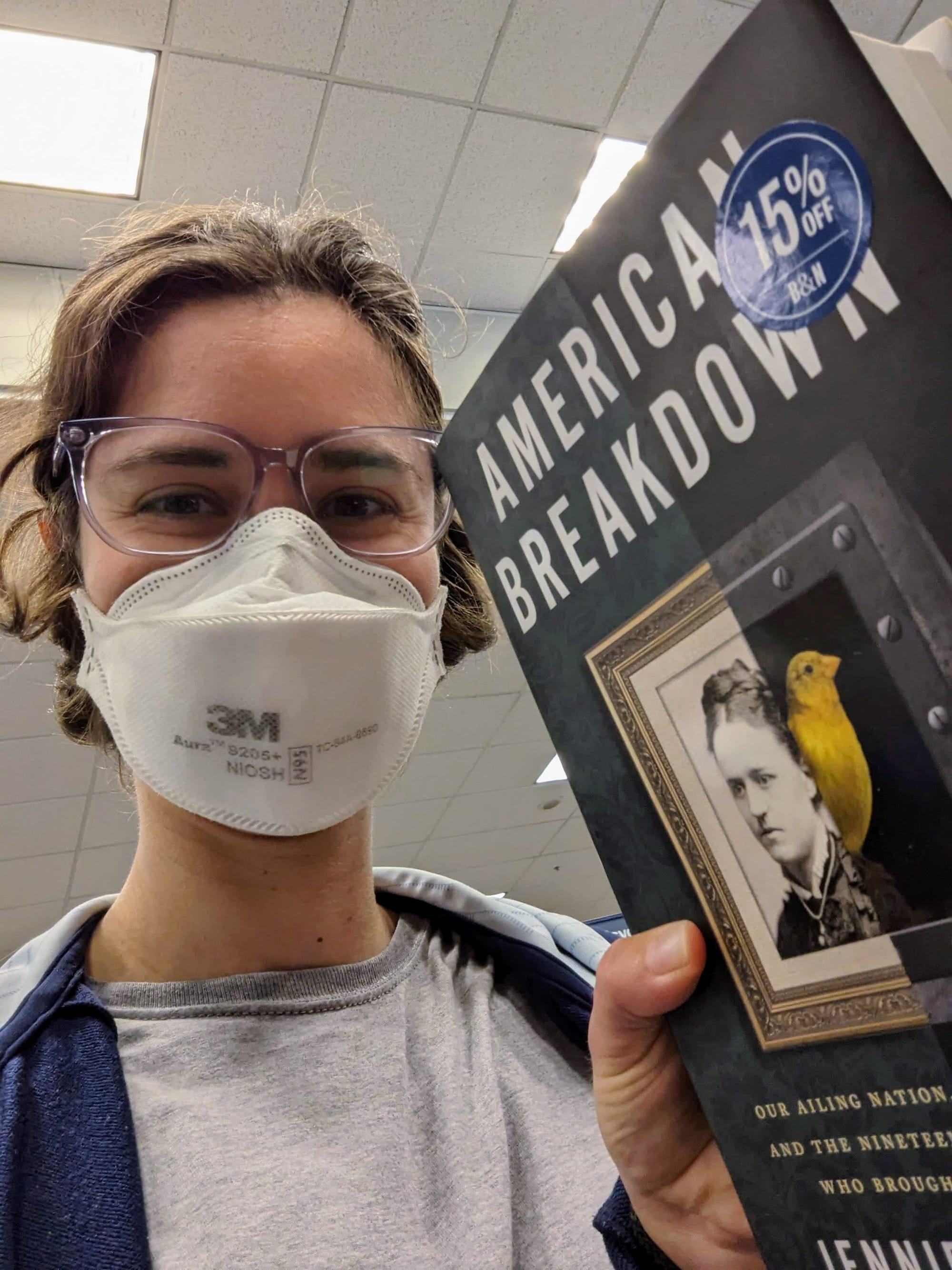 A selfie of Rey wearing a mask in a bookstore, holding a copy of American Breakdown by Jennifer Lunden