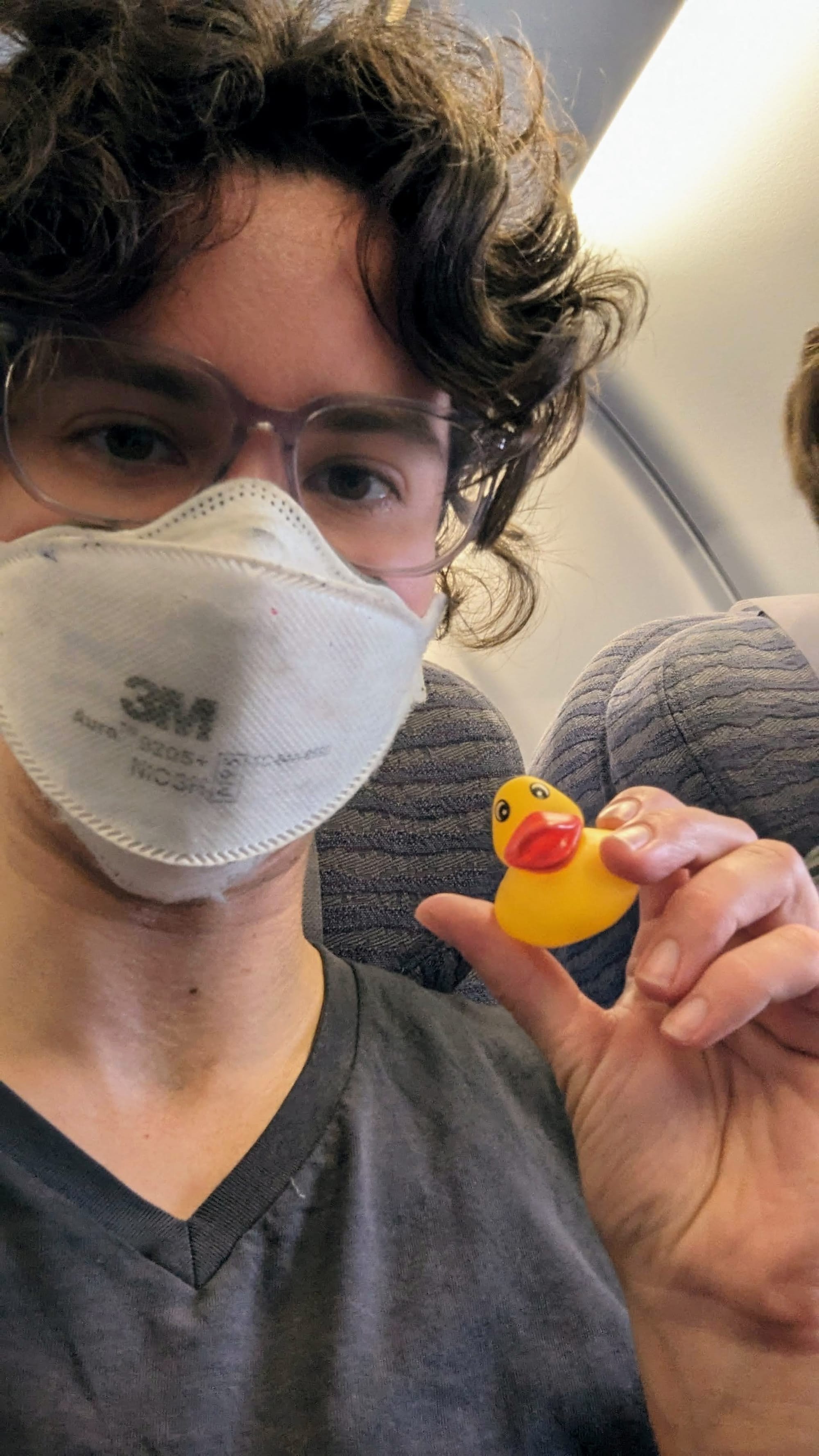 A photo of Rey wearing a mask, sitting in an airplane seat, holding a small, cute rubber duck