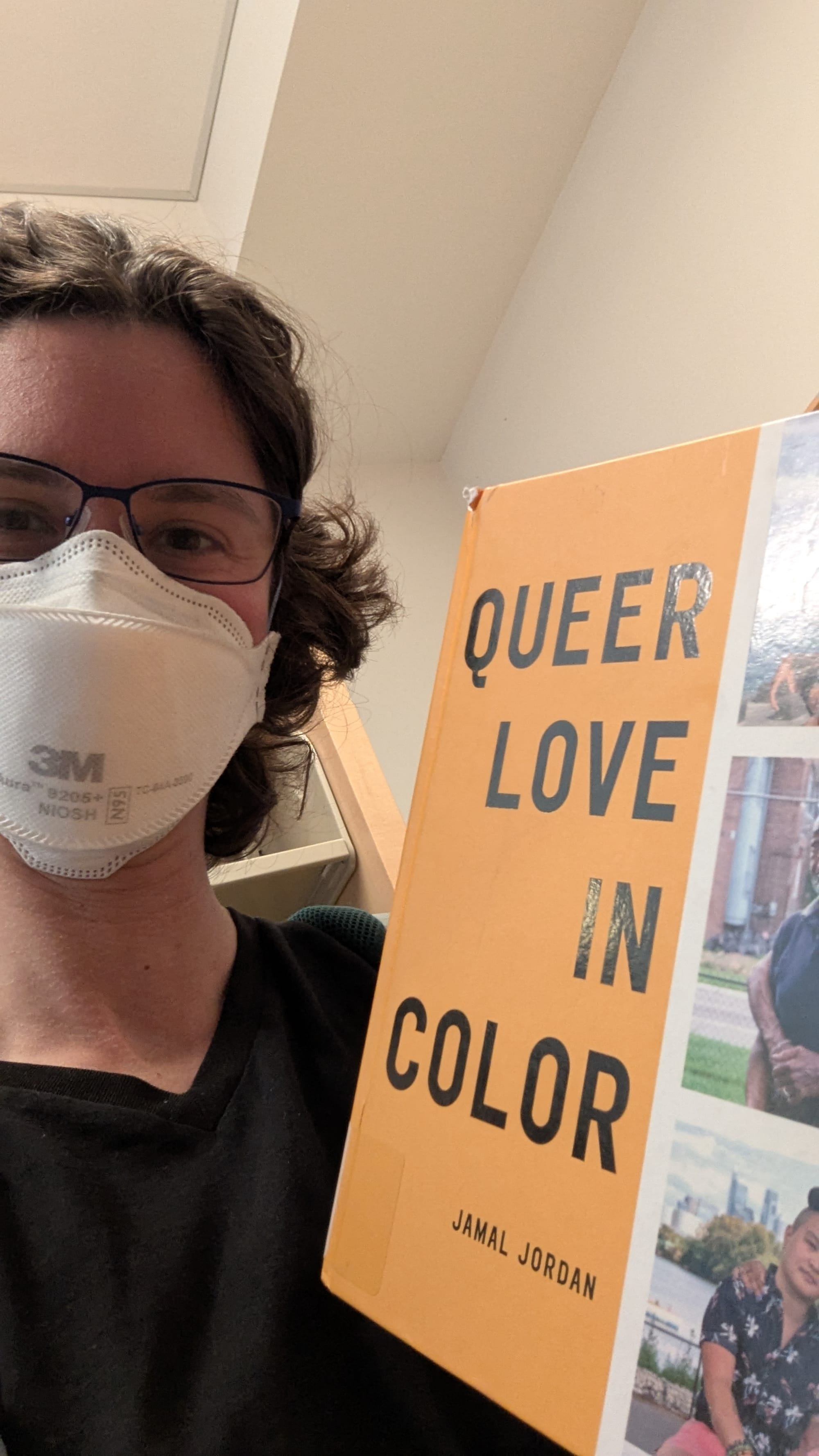 A photo of Rey wearing a mask in a bookstore, holding the book Love in Color by Jamal Jordan